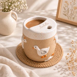 May include: A ceramic mug with a speckled cream top and a tan base, featuring raised white goose figures with orange beaks and feet. The rim includes a cutout for a straw and the name Isabella engraved in cursive script on the top surface.