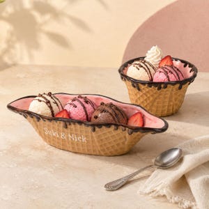May include: A long, oval-shaped ceramic bowl and a round ceramic bowl, both textured like waffle cones with dark brown drips along the rim. The long bowl features the white text Suki & Nick. Both contain scoops of ice cream with chocolate drizzle.