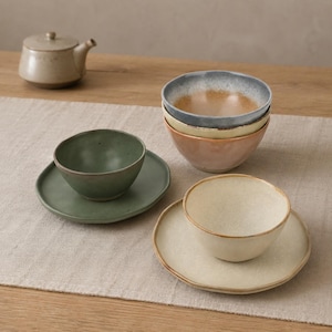 May include: A ceramic tea set featuring a light beige teapot, a green bowl on a matching saucer, a cream-colored bowl on a matching saucer, and a stack of two bowls in blue and orange tones, all with a speckled, matte finish and organic shapes.
