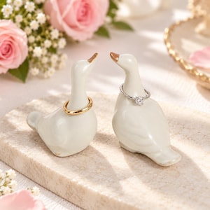 May include: Two white ceramic goose-shaped ring holders with gold-painted beaks. One goose holds a plain gold band around its neck, while the other holds a silver-toned ring featuring a single clear round gemstone setting.