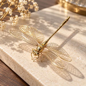 May include: A gold-colored dragonfly figurine featuring intricate, open-work filigree wings with a detailed vein pattern. The body is segmented with a smooth, polished finish, and the head includes two prominent eyes and thin, delicate legs.