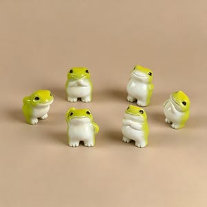 May include: A collection of six small, standing frog figurines. Each frog features a smooth, glossy finish with a bright lime green back and a white belly. The frogs are depicted in various poses, including some with arms crossed or raised, all with black eyes.
