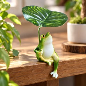 Könnte beinhalten: Eine grün-weiße Froschfigur, die auf einer Kante sitzt und ein großes grünes Blatt wie einen Regenschirm über dem Kopf hält. Der Frosch hat einen glatten, runden Körper, gelb umrandete Augen und ein Bein, das nach unten hängt.