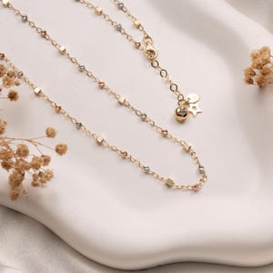 May include: A delicate gold chain necklace featuring alternating small metallic cubes and textured beads. The necklace includes a lobster claw clasp, a short extender chain, and two small dangling charms shaped like a sphere and a star.