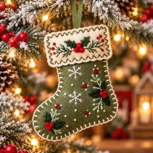 May include: A felt Christmas stocking in olive green with a cream-colored cuff. The stocking features embroidered white snowflakes, green holly leaves, and red berries. It is finished with a decorative cream blanket stitch along the edges.