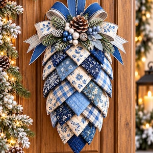 May include: A decorative hanging ornament shaped like a large pinecone, constructed from layered fabric patches in various shades of blue and cream. The fabric features floral, plaid, and solid patterns. It is topped with a blue ribbon bow, pinecones, and berries.