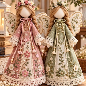 May include: Two fabric angel dolls with braided hair and floral crowns. One doll wears a dusty rose dress, the other a sage green dress. Both feature embroidered wings, lace-trimmed hems, pearl accents, and intricate floral embroidery on the skirts.