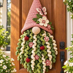 May include: A crocheted gnome wall hanging featuring a dusty pink pointed hat adorned with a white butterfly and cream-colored flowers. The long beard consists of curly green and cream yarn strands decorated with small red ladybugs and pink flowers.