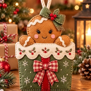 May include: A felt gingerbread person ornament peeking out of a green gift box. The gingerbread figure has a green bow with red berries, while the box features a white scalloped trim, embroidered snowflakes, and a red and white checkered bow.