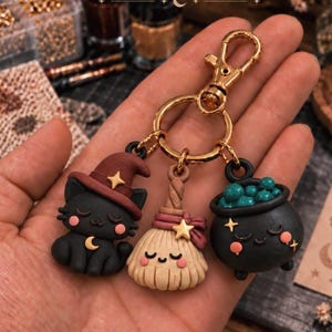 Könnte beinhalten: Ein goldfarbener Schlüsselanhänger mit drei kleinen Anhängern: eine schwarze Katze mit braunem Hexenhut, ein beigefarbener Besen mit rosa Schleife und ein schwarzer Kessel mit grünen Blasen. Jeder Anhänger hat ein lächelndes Gesicht mit rosa Wangen.