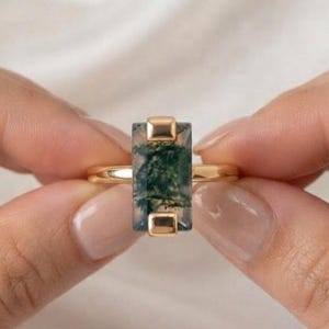 May include: A gold ring featuring a rectangular moss agate gemstone with green inclusions. The stone is held in a vertical setting with gold prongs at the top and bottom, connected to a thin, polished gold band that encircles the finger.