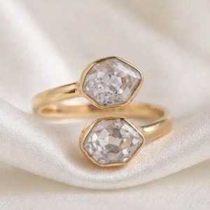 Puede incluir: Un anillo abierto de oro con dos piedras transparentes de corte hexagonal en cada extremo. La banda es de oro pulido y las dos piedras facetadas están colocadas frente a frente con un pequeño espacio, creando un diseño abierto.