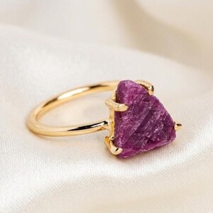 Op de afbeelding: Een gouden ring met een driehoekige, ruwe, diep magentakleurige edelsteen in een klauwzetting. De steen heeft een getextureerd, kristallijn oppervlak en de band is van glad, gepolijst goudkleurig metaal dat het cirkelvormige ontwerp vormt.