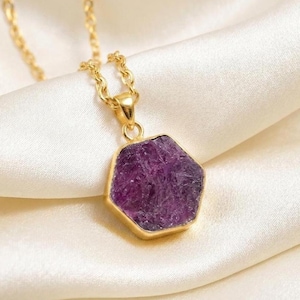 Puede incluir: Un collar de cadena dorada con un colgante hexagonal que presenta una piedra de amatista en bruto de color púrpura intenso. El colgante está engastado en un marco de metal dorado y la cadena consta de pequeños eslabones dorados pulidos.
