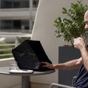 May include: A black laptop privacy hood with the text MNK printed in white on the front edge. The foldable accessory is positioned on a round table next to a clear glass of water and a notebook, designed to reduce screen glare for laptop users.
