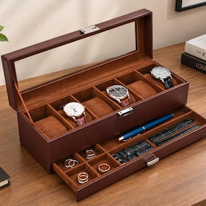 May include: A brown leather watch box with a glass lid, featuring a top compartment with six watch pillows and a pull-out drawer containing four small square jewelry slots, a blue pen, and two black watch straps with silver buckles.