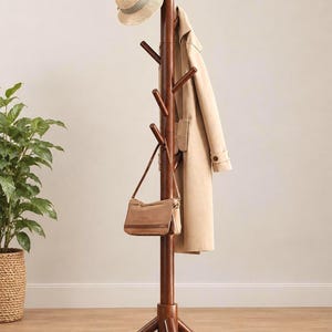 May include: A tall, dark brown wooden coat rack with a central vertical post and multiple angled pegs. The rack holds a beige trench coat, a straw hat with a grey ribbon, and a small brown shoulder bag hanging from one of the wooden hooks.