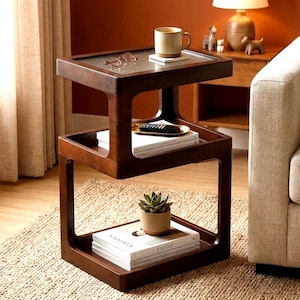 May include: A dark brown wooden side table with three tiers. The top tier features a glass insert, a ceramic mug, and eyeglasses. The middle tier holds a remote control on a small tray and books. The bottom tier holds a potted succulent and books.