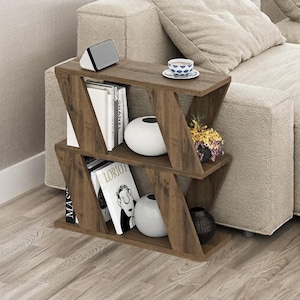 May include: A brown wood-grain side table with a Z-shaped frame featuring two open shelves. The top shelf holds a digital clock, a patterned teacup on a saucer, and a small plant. The bottom shelf contains books, including one titled LORIOT, and two vases.