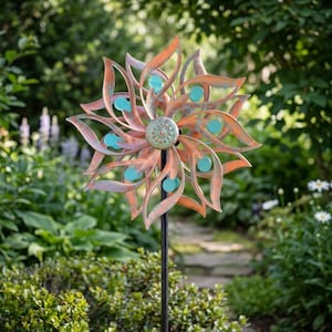 May include: A metal garden wind spinner featuring a multi-layered flower design with copper-toned petals and small, circular turquoise accents. The spinner is mounted on a thin, dark vertical pole, showcasing a symmetrical, rotating structure.