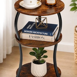 May include: A three-tier round side table with a dark wood grain finish and black metal curved legs. The top shelf holds a white Orion & Co coffee cup and a Cedarwood & Bergamot candle. The middle shelf holds Modern Architecture and Fashion books.