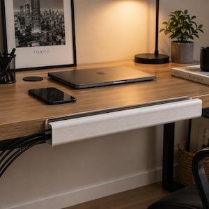 Op de afbeelding: Een wit rechthoekig kabelgootje bevestigd aan de voorrand van een houten bureau, voorzien van een horizontale gleuf die meerdere zwarte stroomkabels ordent en aan de zijkant van de unit verbergt.