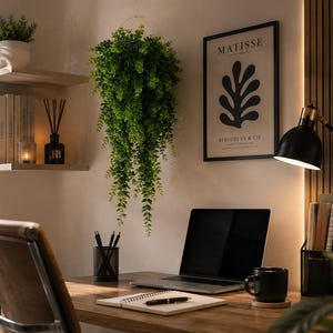 May include: A home office setup featuring a hanging artificial green plant, a framed Matisse Berggruen & Cie print, a laptop, a black desk lamp, a notebook with a pen, a black pencil holder, and a black mug on a wooden desk with wall shelving.