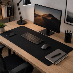 Peut inclure: Un grand tapis de bureau rectangulaire noir recouvrant une surface en bois clair. Le tapis supporte un clavier sans fil, une souris, un smartphone, un carnet gris avec un stylo noir et une tasse noire avec le texte "BUT FIRST COFFEE".