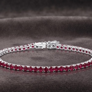 Pode incluir: Uma pulseira de ténis em tom prateado com uma fila contínua de rubis redondos vermelho-escuros engastados em garras de metal. A pulseira inclui um fecho de caixa seguro com trinco de segurança, formando um círculo com pedras uniformes.