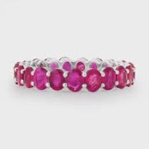 Può includere: Un anello a fascia eternity con una fila continua di rubini rosso-rosa vivaci dal taglio ovale, incastonati in una banda di metallo color argento lucido. Le pietre sono uniformi per dimensioni e forma, creando un design circolare coerente.