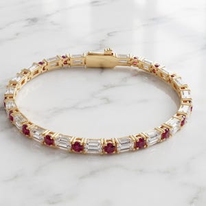 May include: A gold tennis bracelet featuring an alternating pattern of baguette-cut clear stones and round-cut red stones. The bracelet is secured with a gold box clasp closure, creating a continuous circular design of metal and gemstones.