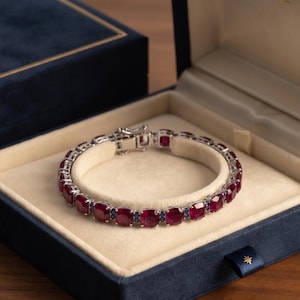 May include: A silver-toned tennis bracelet featuring a series of cushion-cut red rubies alternating with small round blue sapphires. The bracelet has a secure metal clasp and is presented inside a navy blue velvet-lined jewelry display box.