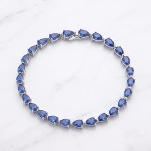Può includere: Un bracciale tennis color argento caratterizzato da una fila continua di pietre preziose blu a taglio pera incastonate in montature di metallo. Il bracciale è disposto in forma circolare e presenta una chiusura metallica sicura in alto.