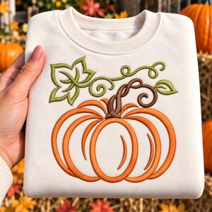May include: A cream-coloured crewneck sweatshirt featuring a large, centred machine embroidery design of an orange pumpkin with a brown stem and green vine leaves. The pumpkin is outlined in a bold, dark orange thread with visible stitching.