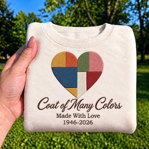 Può includere: Una felpa girocollo color crema con un cuore ricamato composto da segmenti geometrici multicolori in giallo, arancione, blu, verde, rosa, rosso e bianco. Sotto il cuore, il testo marrone recita Coat of Many Colors, Made With Love, 1946-2026.