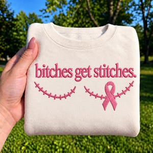May include: A cream-colored crewneck sweatshirt featuring pink embroidered text that reads "bitches get stitches." Below the text are two curved lines of pink embroidered stitches, with the right side incorporating a pink awareness ribbon.