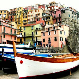 Puede incluir: Un edificio colorido de varios niveles con una fila de barcos en primer plano. Los barcos son blancos con molduras de madera y detalles azules. El edificio est&aacute; construido en un acantilado rocoso.