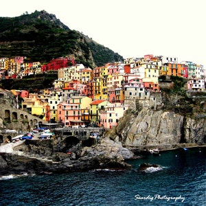 May include: A colourful village built into a cliffside overlooking a blue body of water. The buildings are painted in shades of yellow, orange, red, and green. The village is surrounded by lush green vegetation.