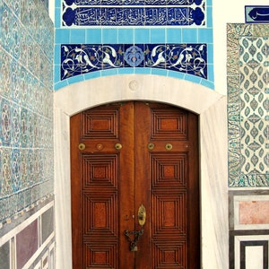 May include: A wooden door with intricate carvings and a metal lock and chain. The door is framed by a white archway with blue and white tile accents. The tiles feature Arabic calligraphy.
