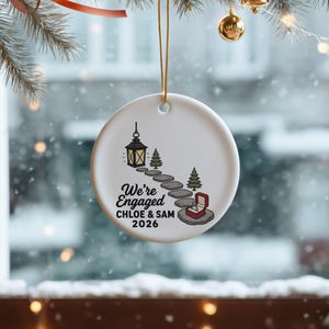 May include: A round white ceramic ornament hanging by a gold string. The design features a black lantern, a stone path, two green pine trees, and an open red ring box. Text reads: We're Engaged, CHLOE & SAM, 2026, in black lettering.