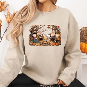 Könnte beinhalten: Ein beigefarbenes Sweatshirt mit Rundhalsausschnitt und einem Grafikdruck der Peanuts-Figuren, die um ein Lagerfeuer in einem Herbstwald sitzen. Die Illustration zeigt Charlie Brown, Snoopy und Freunde mit Kürbissen, Bäumen und einem Kessel.