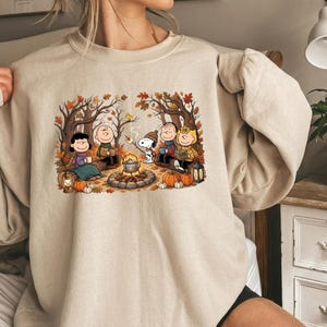 Könnte beinhalten: Ein beigefarbenes Sweatshirt mit Rundhalsausschnitt und einem Grafikdruck der Peanuts-Figuren, die in einem herbstlichen Wald um ein Lagerfeuer sitzen. Die Illustration zeigt Snoopy, Charlie Brown und Freunde mit Kürbissen, Laternen und einem Kessel.