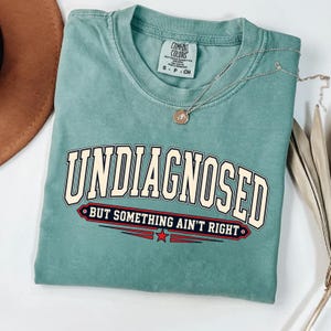 Könnte beinhalten: Ein salbeigrünes Kurzarm-T-Shirt mit dem Text UNDIAGNOSED in einer großen cremefarbenen College-Schriftart und dem darunter gedruckten Satz BUT SOMETHING AIN'T RIGHT in einem rot-dunkelblauen Banner-Design mit einem kleinen roten Stern.