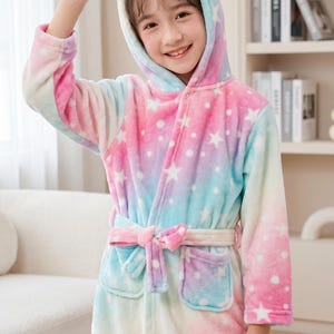 May include: A plush, long-sleeve bathrobe featuring a pastel tie-dye pattern of pink, blue, and white with a white star print. The robe includes a hood with a unicorn horn and ears, a tie-waist belt, and two front pockets made of soft fleece.