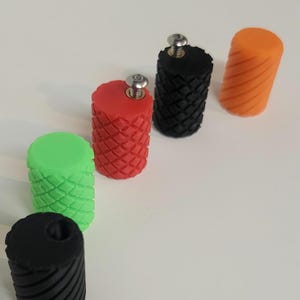 May include: A collection of six cylindrical knobs in various colors including black, lime green, red, and orange. Each knob features a textured surface with either a diamond or diagonal pattern. Two of the knobs have silver metal screws inserted.