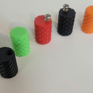 May include: A collection of cylindrical knobs in various colors including black, lime green, red, and orange. The knobs feature textured surfaces with either a diamond or diagonal ribbed pattern. Some knobs include a silver metal screw at the top.