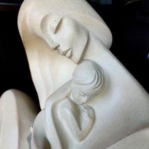May include: A cream-colored sculpture featuring a stylized figure embracing a smaller figure. The material has a speckled texture throughout, with smooth, flowing lines defining the faces, hair, and bodies of the two figures in a close, protective pose.