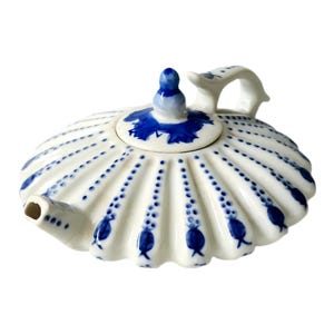 May include: A white ceramic teapot with a shell-like design, adorned with blue floral patterns and dotted accents. The lid features a blue finial and matching blue floral details. The handle is curved and integrated into the design.