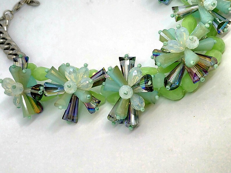 May include: A green and iridescent crystal beaded necklace with a silver chain. The beads are arranged in a flower shape.