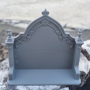 Pode incluir: Um suporte de exibição cinza impresso em 3D com um painel traseiro arqueado com volutas decorativas e dois remates nos pilares laterais. A estrutura tem uma base plana e um acabamento fosco liso, projetado para segurar pequenos objetos.